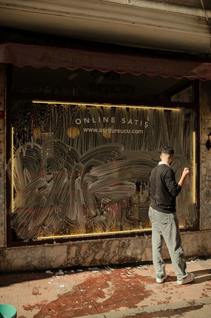 A man wearing a black jacket, light-colored trousers, and glasses is cleaning a large glass storefront window at Alexandra Palace, possibly using a cloth or spray. The window features graffiti-style artwork with swirling patterns and text reading 'ONLINE SATIS'. Behind the glass, there is a dimly lit interior with visible shelving and merchandise. The exterior sidewalk appears slightly dirty with patches of debris, and the scene is illuminated by natural daylight. The overall setting reflects a deep cleaning or maintenance process carried out by Deep Cleaning Haringey, ensuring the surface remains clear and hygienic for customers and passersby.