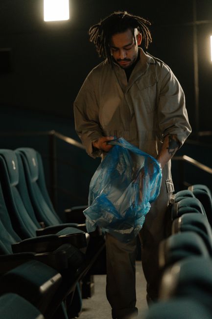 A male cleaner with dreadlocks, wearing a beige uniform, is performing surface cleaning in an auditorium or theater setting. He is holding a blue plastic bag, possibly for waste collection or disposal of cleaning materials. The background features rows of dark teal upholstered chairs with metal armrests, arranged in a tiered formation. The polished wooden or laminate flooring reflects light, indicating a well-maintained and sanitized environment. Bright overhead lighting illuminates the space, emphasizing the cleanliness and care being taken during the deep cleaning process. This scene illustrates professional commercial cleaning, emphasizing hygiene and maintenance in a public venue, consistent with services offered by Deep Cleaning Haringey at Alexandra Palace event spaces.