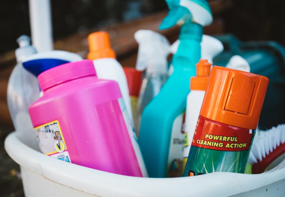 A white plastic bucket containing various cleaning products and tools for surface cleaning and sanitation. Visible items include a large pink liquid detergent bottle, an orange aerosol can with a label stating 'Powerful Cleaning Action', several spray bottles in white, teal, and blue with trigger nozzles, and a red cleaning brush. The bottles are arranged vertically with some leaning against each other, and the scene appears to be outdoors with natural lighting illuminating the bottles. The cleaning supplies are clean and organized, ready for domestic deep cleaning tasks. This image reflects the range of products used by Deep Cleaning Haringey in their residential and commercial cleaning services, particularly for maintaining hygiene and surface cleanliness, fitting with the focus of the page on deep cleaning tips for local landlords in Broadwater Farm, Haringey.