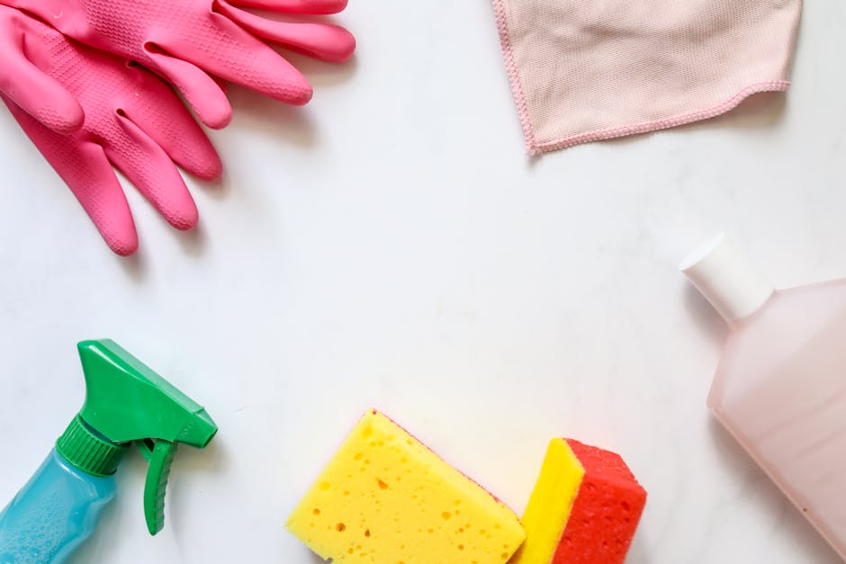 A flat lay of cleaning tools and materials on a white surface, including a pair of pink rubber gloves, a pink cleaning cloth, a green spray bottle, a yellow and red cleaning sponge, and a white plastic bottle with a white cap, arranged for surface cleaning or deep cleaning tasks. The items are clean and ready for domestic or commercial sanitation, reflecting thorough hygiene practices as recommended by Deep Cleaning Haringey for local landlords in Broadwater Farm, Haringey.