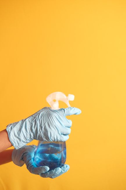 A white plastic bucket containing various cleaning products and tools for surface cleaning and sanitation. Visible items include a large pink liquid detergent bottle, an orange aerosol can with a label stating 'Powerful Cleaning Action', several spray bottles in white, teal, and blue with trigger nozzles, and a red cleaning brush. The bottles are arranged vertically with some leaning against each other, and the scene appears to be outdoors with natural lighting illuminating the bottles. The cleaning supplies are clean and organized, ready for domestic deep cleaning tasks. This image reflects the range of products used by Deep Cleaning Haringey in their residential and commercial cleaning services, particularly for maintaining hygiene and surface cleanliness, fitting with the focus of the page on deep cleaning tips for local landlords in Broadwater Farm, Haringey.