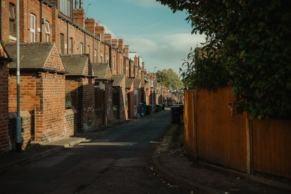 A narrow residential alleyway in Haringey featuring a row of brick terraced houses with small front yards, bordered by a wooden fence on the right side and a brick wall with small roofed structures ov